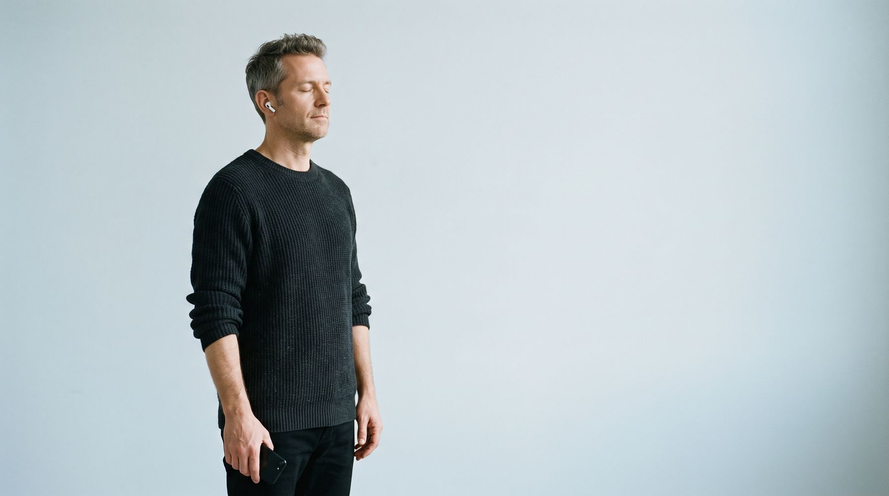 A man standing in a pale studio with his eyes closed, one earbud in, listening