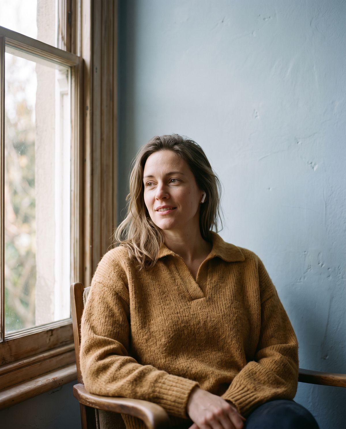A woman by a window, earbuds in, listening with a faint smile